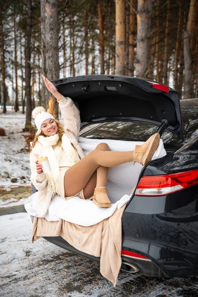 Young woman in woolen hat sits in the trunk of the car. Girl have a fun in black car. Winter travel, snow-covered trees in the forest. Snowy weather. . High quality photo