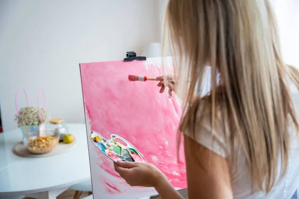 Young woman artist with palette and brush painting abstract pink picture on canvas at home. Back view. Art and creativity concept. High quality photo