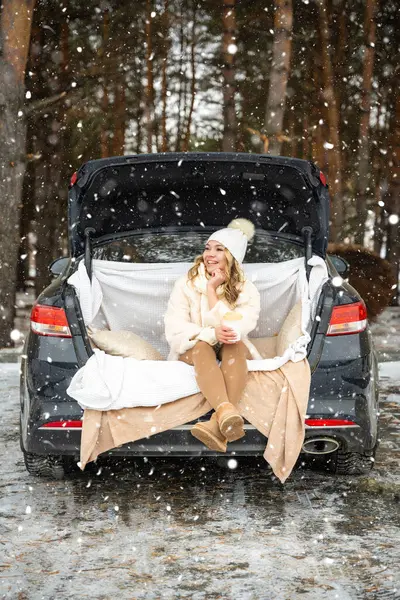 Young woman in woolen hat sits in the trunk of the car. Girl have a fun in the car. Winter travel in the forest. Snowy weather. High quality photo