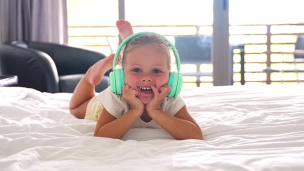 小さな女の子は 家庭のベッドで緑の子供のヘッドフォンで音楽を楽しんでいます 高品質の4K映像 — ストック動画