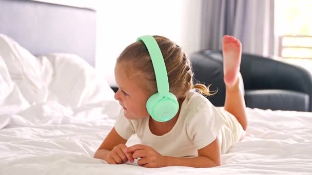 Маленька Дівчинка Лежить Ліжку Слухає Музику Зеленими Дитячими Навушниками Домашній — стокове відео