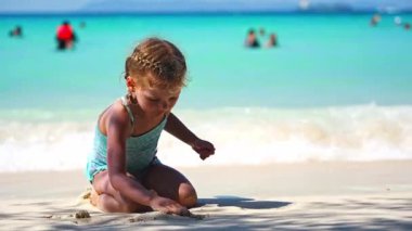 Denizdeki kumda oynayan küçük bir kız, deniz beyazının arka planı ve mavi su. Tatil ve çocuk konseptiyle seyahat. Yüksek kalite 4k görüntü