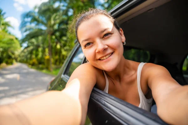 Happy young woman with dark hair stuck her hands out of the cab of a car and make selfie on the road, concept of auto travel. High quality photo