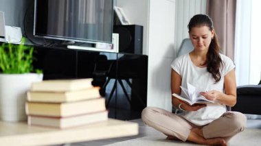 Genç kadın oturma odasında yerde oturmuş kitap okuyor, ön planda kitap yığını. Yüksek kalite 4k görüntü