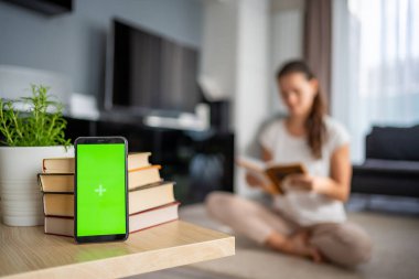 Digital detox concept photo. Smartphone with green chroma key screen and woman reading book in the background. High quality photo clipart