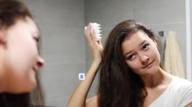 Koyu kıvırcık saçlı genç bir kadın kafa derisi masajı yapıyor ya da saç büyümesi için saç fırçasıyla ev banyosunda masaj yapıyor. Aynanın yansıyan görüntüsü. Yüksek kalite fotoğraf