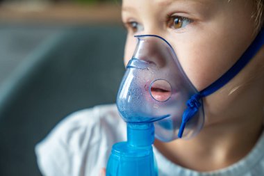 Küçük kızın yakın plan görüntüsü nebulizör maskesini tutuyor ve yüzüne yaslanıyor, solunum yolu tedavisi kavramı. Yüksek kalite fotoğraf