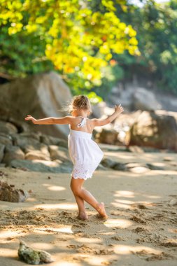 Beyaz elbiseli küçük kız ağaçların ve palmiyelerin gölgesinde sahilde eğleniyor. Yüksek kalite fotoğraf