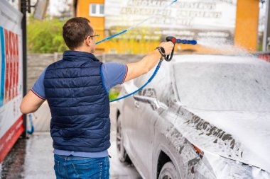 Genç bir adam arabasını tazyikli su ve köpüklü bir hortumla oto yıkamada yıkıyor. Yüksek kalite fotoğraf
