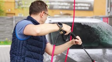 Genç bir adam arabasını tazyikli su ve köpüklü bir hortumla oto yıkamada yıkıyor. Yüksek kalite 4k görüntü