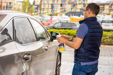 Genç adam oto yıkamada arabanın camlarını temizlemek için sıvı kullanıyor.