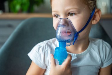 Küçük kızın yakın plan görüntüsü nebulizör maskesini tutuyor ve yüzüne yaslanıyor, solunum yolu tedavisi kavramı. Yüksek kalite fotoğraf