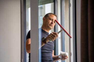 Genç adam evdeki pencereleri temizler. Deterjan sıkar, bardağa su sürer ve temizler. Yüksek kalite fotoğraf
