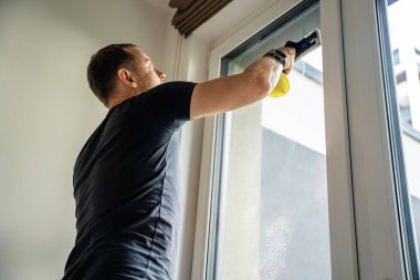 Genç adam evdeki pencereleri temizler. Deterjan sıkar, bardağa su sürer ve temizler. Yüksek kalite fotoğraf
