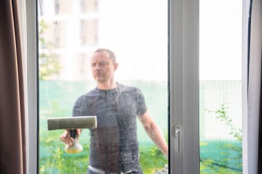 Genç adam evdeki pencereleri temizler. Deterjan sıkar, bardağa su sürer ve temizler. Yüksek kalite fotoğraf