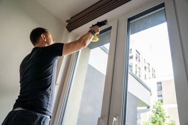 Genç adam evdeki pencereleri temizler. Deterjan sıkar, bardağa su sürer ve temizler. Yüksek kalite fotoğraf