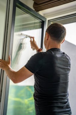 Genç adam evdeki pencereleri temizler. Deterjan sıkar, bardağa su sürer ve temizler. Yüksek kalite fotoğraf