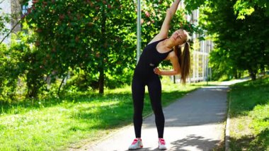 Siyah spor giysili genç bir kadın şehir parkında egzersiz yapıyor. Koşmadan ya da koşmadan önce ısın. Açık hava sporları eğitimi konsepti. Yüksek kalite 4k görüntü