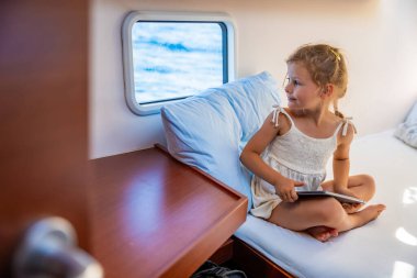 Küçük kız elinde tabletle bir yatın kabininde oturur ve bir deniz geçidi sırasında pencereden dışarı bakar. Yat hayatı konsepti. Yüksek kalite fotoğraf