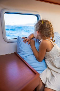 Sarışın küçük kız bir yatın kabininde oturur ve bir deniz geçişi sırasında pencereden dışarı bakar. Yat hayatı konsepti. Yüksek kalite fotoğraf