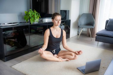 Spor kıyafetli genç bir kadın oturma odasında minder üzerinde yoga egzersizi yapıyor. Çevrimiçi egzersiz, fitness, pratik ve evde meditasyon. Yüksek kalite fotoğraf