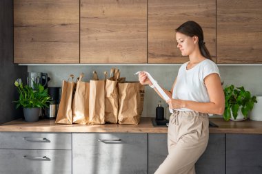 Mutfakta içi yiyecek dolu kağıt torbaların yanındaki dükkandan alınmış fişi olan genç bir kadın. Yiyecekler internetten sipariş edildi ve kuryeyle teslim edildi. Yüksek kalite fotoğraf