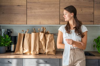 Mutfakta içi yiyecek dolu kağıt torbaların yanındaki dükkandan alınmış fişi olan genç bir kadın. Yiyecekler internetten sipariş edildi ve kuryeyle teslim edildi. Yüksek kalite fotoğraf