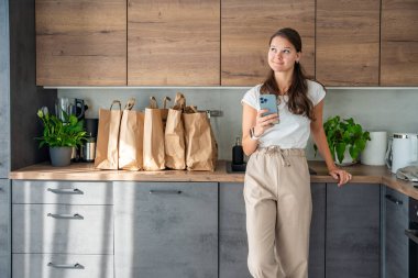 Elinde akıllı telefon olan genç bir kadın mutfakta içi yiyecek dolu kağıt torbaların yanında. Yiyecekler internetten sipariş edildi ve kuryeyle teslim edildi. Yüksek kalite fotoğraf