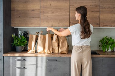 Mutfakta yiyecek dolu kağıt torbaların yanındaki genç kadın. Yiyecekler internetten sipariş edildi ve kuryeyle teslim edildi. Yüksek kalite fotoğraf
