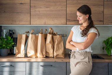 Mutfakta yiyecek dolu kağıt torbaların yanındaki genç kadın. Yiyecekler internetten sipariş edildi ve kuryeyle teslim edildi. Yüksek kalite fotoğraf