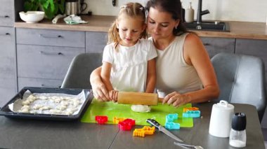 Küçük kız, annesinin hamuru hamurlu kurabiye yapmak ve mutfak mutfağında küflü kurabiye makası yapmak için ahşap hamurla yuvarlamasına yardım ediyor. Mutfakta mutlu bir aile. Yüksek kalite 4k görüntü