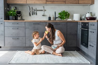 Anne ve kızı mutfak zemininde oturuyor, yemek yiyor ya da kahvaltı edip oynuyorlar. Aile bağı. Yüksek kalite fotoğraf