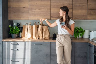 Elinde akıllı telefon olan genç bir kadın mutfakta içi yiyecek dolu kağıt torbaların yanında. Yiyecekler internetten sipariş edildi ve kuryeyle teslim edildi. Yüksek kalite fotoğraf