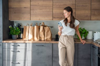 Elinde akıllı telefon olan genç bir kadın mutfakta içi yiyecek dolu kağıt torbaların yanında. Yiyecekler internetten sipariş edildi ve kuryeyle teslim edildi. Yüksek kalite fotoğraf