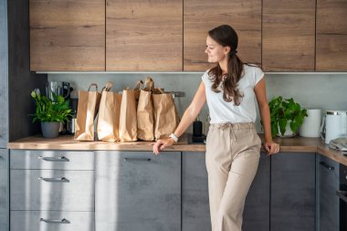 Mutfakta yiyecek dolu kağıt torbaların yanındaki genç kadın. Yiyecekler internetten sipariş edildi ve kuryeyle teslim edildi. Yüksek kalite fotoğraf