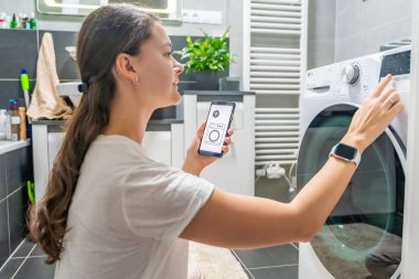 Genç bir kadın cep telefonu kontrolünü kablosuz ağ ve uygulamayla evde çamaşır makinesi olarak kullanıyor. Homesmart ve yeni teknoloji konsepti. Yüksek kalite fotoğraf