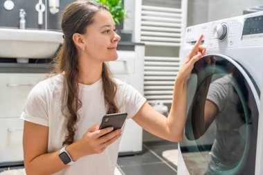 Genç bir kadın cep telefonu kontrolünü kablosuz ağ ve uygulamayla evde çamaşır makinesi olarak kullanıyor. Homesmart ve yeni teknoloji konsepti. Yüksek kalite fotoğraf
