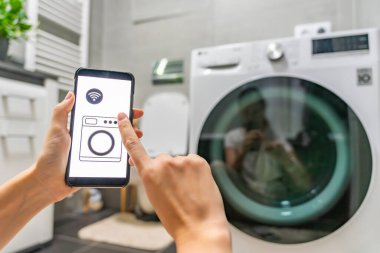 Genç bir kadın cep telefonu kontrolünü kablosuz ağ ve uygulamayla evde çamaşır makinesi olarak kullanıyor. Homesmart ve yeni teknoloji konsepti. Yüksek kalite fotoğraf