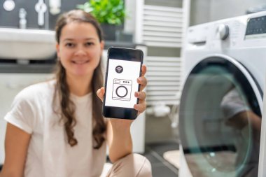 Genç bir kadın cep telefonu kontrolünü kablosuz ağ ve uygulamayla evde çamaşır makinesi olarak kullanıyor. Homesmart ve yeni teknoloji konsepti. Yüksek kalite fotoğraf
