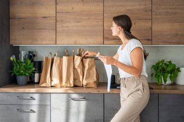 Mutfakta içi yiyecek dolu kağıt torbaların yanındaki dükkandan alınmış fişi olan genç bir kadın. Yiyecekler internetten sipariş edildi ve kuryeyle teslim edildi. Yüksek kalite fotoğraf