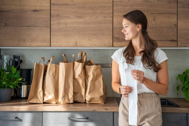 Mutfakta içi yiyecek dolu kağıt torbaların yanındaki dükkandan alınmış fişi olan genç bir kadın. Yiyecekler internetten sipariş edildi ve kuryeyle teslim edildi. Yüksek kalite fotoğraf