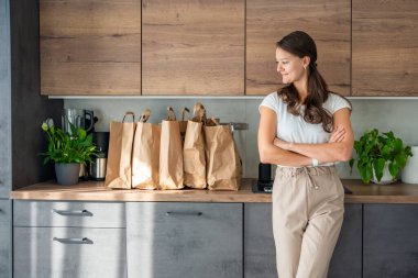 Mutfakta yiyecek dolu kağıt torbaların yanındaki genç kadın. Yiyecekler internetten sipariş edildi ve kuryeyle teslim edildi. Yüksek kalite fotoğraf