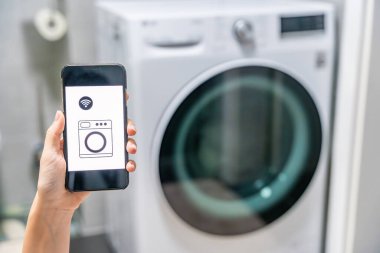 Genç bir kadın cep telefonu kontrolünü kablosuz ağ ve uygulamayla evde çamaşır makinesi olarak kullanıyor. Homesmart ve yeni teknoloji konsepti. Yüksek kalite fotoğraf