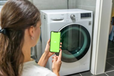 Genç bir kadın cep telefonu kontrolünü kablosuz ağ ve uygulamayla evde çamaşır makinesi olarak kullanıyor. Homesmart uygulaması, krom anahtar yeşil ekran. Yüksek kalite fotoğraf