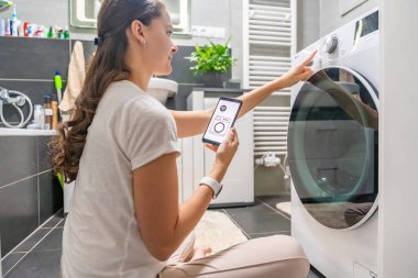 Genç bir kadın cep telefonu kontrolünü kablosuz ağ ve uygulamayla evde çamaşır makinesi olarak kullanıyor. Homesmart ve yeni teknoloji konsepti. Yüksek kalite fotoğraf