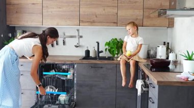 Bir kadın, kızı yanında otururken temiz seramik tabakları bulaşık makinesinden çıkarır. Ev ve kullanışlı teknoloji konsepti. Yüksek kalite 4k görüntü