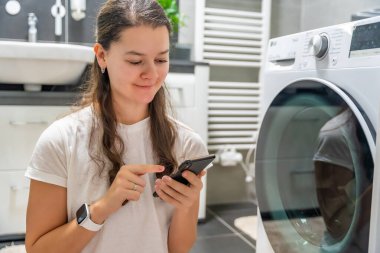 Genç bir kadın cep telefonu kontrolünü kablosuz ağ ve uygulamayla evde çamaşır makinesi olarak kullanıyor. Homesmart ve yeni teknoloji konsepti. Yüksek kalite fotoğraf