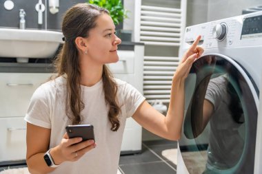 Genç bir kadın cep telefonu kontrolünü kablosuz ağ ve uygulamayla evde çamaşır makinesi olarak kullanıyor. Homesmart ve yeni teknoloji konsepti. Yüksek kalite fotoğraf