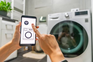Genç bir kadın cep telefonu kontrolünü kablosuz ağ ve uygulamayla evde çamaşır makinesi olarak kullanıyor. Homesmart ve yeni teknoloji konsepti. Yüksek kalite fotoğraf