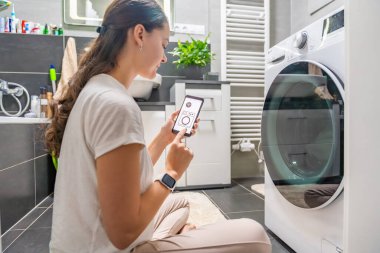 Genç bir kadın cep telefonu kontrolünü kablosuz ağ ve uygulamayla evde çamaşır makinesi olarak kullanıyor. Homesmart ve yeni teknoloji konsepti. Yüksek kalite fotoğraf